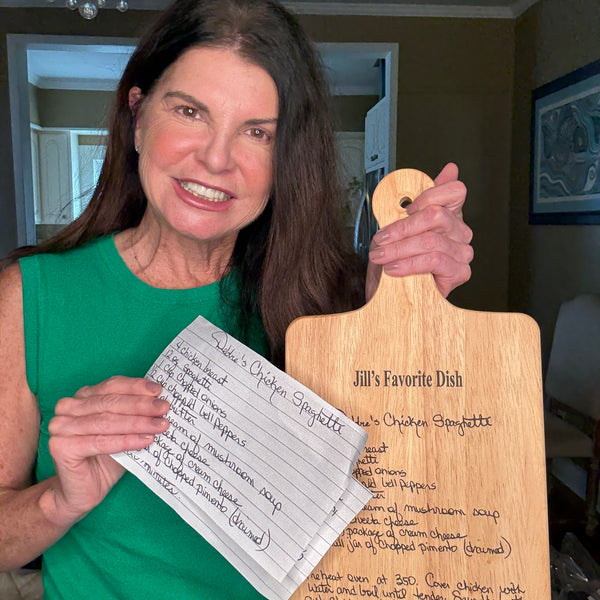 Woman holding a wooden cutting board with a recipe on it, standing in a home setting.