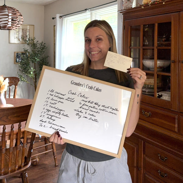 Woman holding a framed recipe in a home setting with wooden furniture and decor.