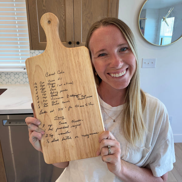 Woman holding a wooden cutting board with a printed recipe in a kitchen.