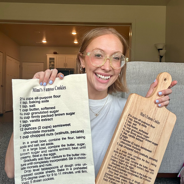 Person holding a recipe tea towel and wooden family recipe cutting board with 'Mom's Famous Cookies' recipe.