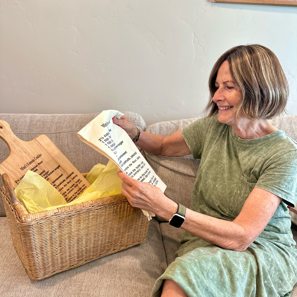 Woman sitting on a couch holding a recipe tea towel bag with a recipe printed on a cutting board next to her. The Printed Gift